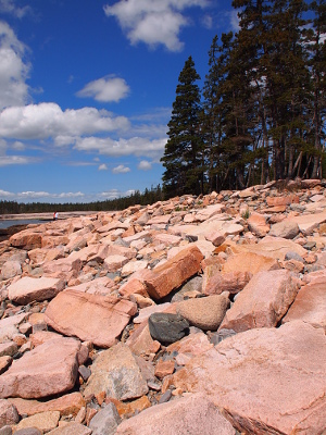 [Granite chunks (1-2 feet in size) littering the shoreline with tall everygreens at the shore and puffy white clouds in the blue sky.]